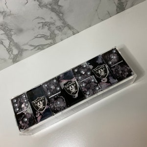 May include: A set of black and white dominoes with the Las Vegas Raiders logo on each tile. The dominoes are in a clear acrylic case.