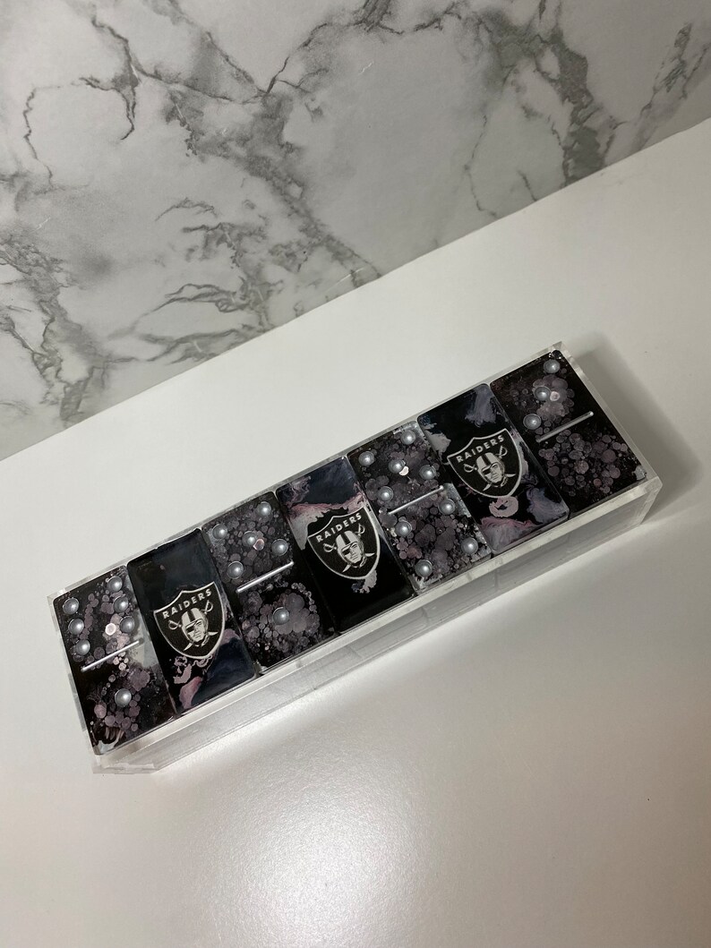 May include: A set of black and white dominoes with the Las Vegas Raiders logo on each tile. The dominoes are in a clear acrylic case.