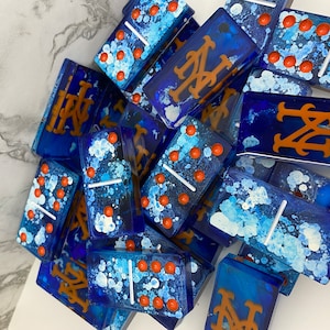 May include: A set of blue dominoes with white and orange paint splatters. Some of the dominoes have the New York Mets logo in orange.