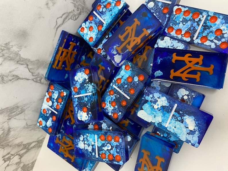 May include: A set of blue dominoes with white and orange paint splatters. Some of the dominoes have the New York Mets logo in orange.