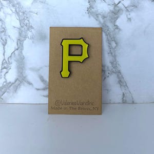 May include: A yellow letter P pin with a black outline attached to a rectangular brown card. The card features the text @ValeriesVianInc and Made in The Bronx, NY printed in black ink at the bottom of the display card.