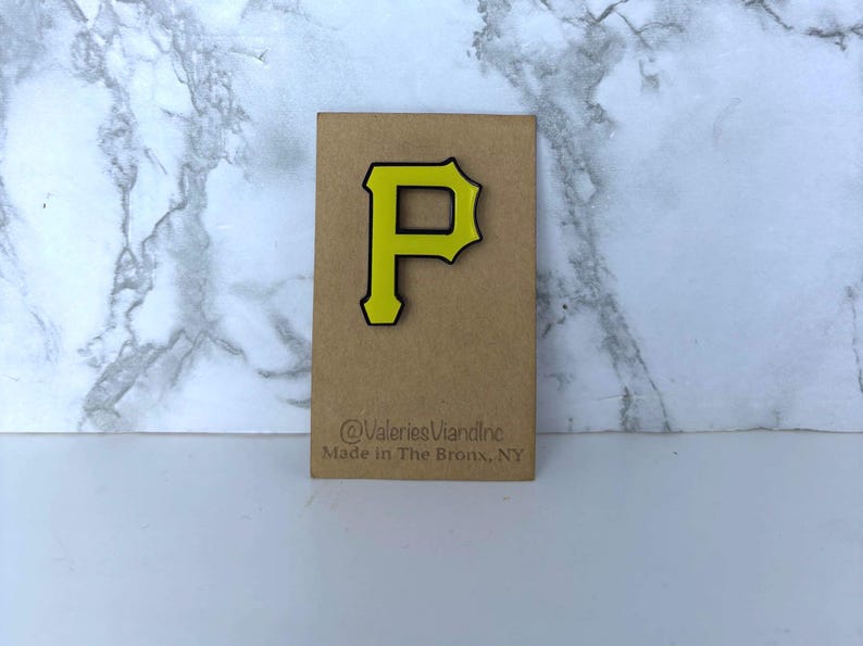 May include: A yellow letter P pin with a black outline attached to a rectangular brown card. The card features the text @ValeriesVianInc and Made in The Bronx, NY printed in black ink at the bottom of the display card.