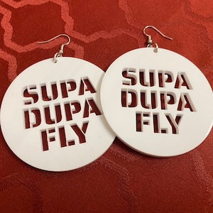 May include: A pair of white circular wooden earrings with the words "Supa Dupa Fly" cut out in the center.