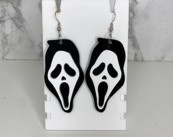 May include: A pair of dangling earrings featuring a black and white ghost face design. The earrings are made of a flat, smooth material and hang from silver-toned metal hooks. The iconic mask shape is clearly defined with black outlines.
