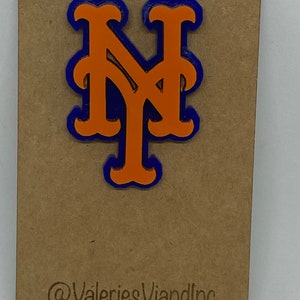 May include: A layered orange and blue NY logo pin attached to a rectangular brown card. The card features the text @ValeriesViandInc and Made in The Bronx, NY printed in black at the bottom. The pin has a bold, stylized design with orange letters.