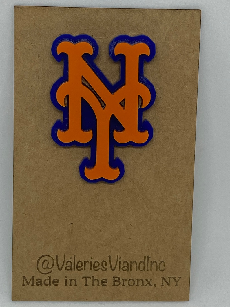 May include: A layered orange and blue NY logo pin attached to a rectangular brown card. The card features the text @ValeriesViandInc and Made in The Bronx, NY printed in black at the bottom. The pin has a bold, stylized design with orange letters.