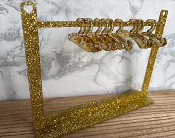 May include: A gold glitter acrylic display rack featuring two vertical side supports with heart-shaped cutouts at the top, a horizontal crossbar, and a rectangular base. Six small matching gold glitter acrylic hangers hang from the crossbar.