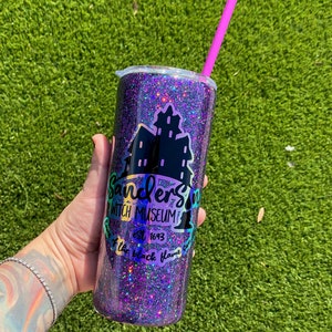 Puede incluir: Un vaso de purpurina morado con un logotipo negro que dice "Sandersons Witch Museum est 1693 Keep the black flame burning".