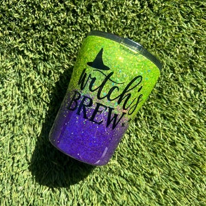 May include: A green and purple glitter tumbler with a black witch's hat and the text "witch's brew".