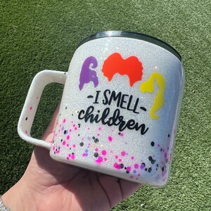 May include: White glitter tumbler with a black lid and handle. The tumbler has a black and white graphic of three witches with the text "I smell children".
