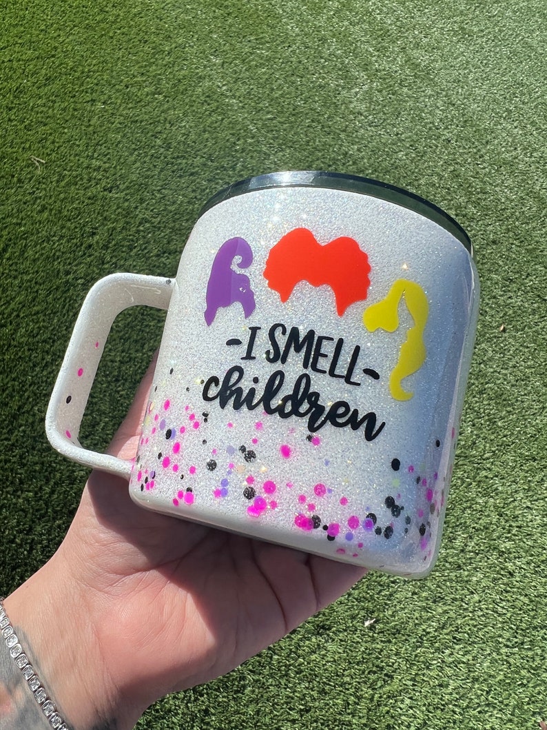 May include: White glitter tumbler with a black lid and handle. The tumbler has a black and white graphic of three witches with the text "I smell children".