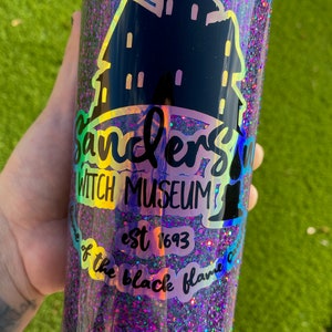 Puede incluir: Un vaso de glitter negro y morado con una silueta negra de una casa y el texto "Sanders Witch Museum est 1693 home of the black flame candle".