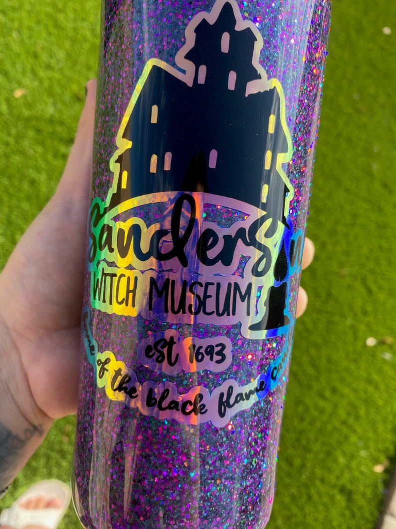 Puede incluir: Un vaso de glitter negro y morado con una silueta negra de una casa y el texto "Sanders Witch Museum est 1693 home of the black flame candle".