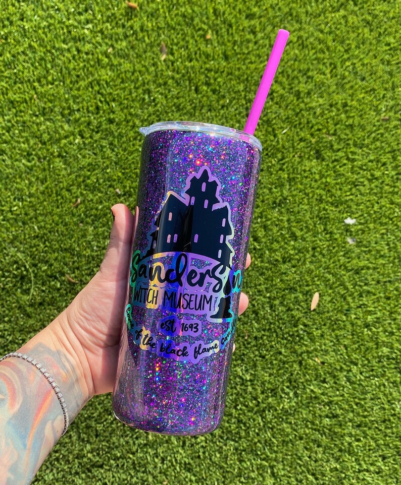 Puede incluir: Un vaso de purpurina morado con un logotipo negro que dice "Sandersons Witch Museum est 1693 Keep the black flame burning".