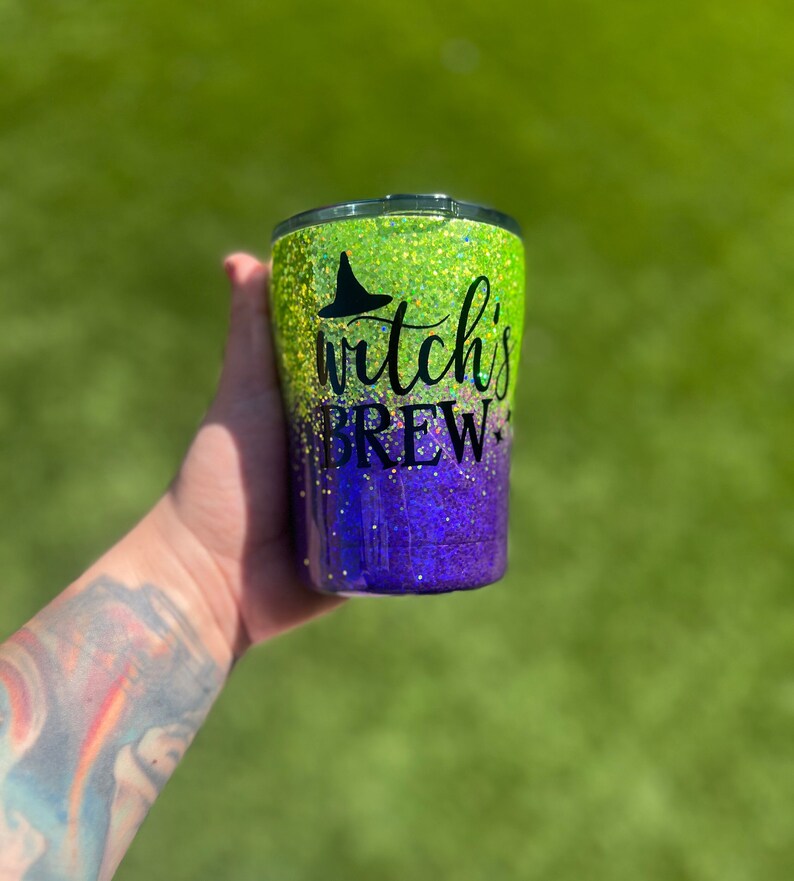 May include: A green and purple glitter tumbler with a black witch's hat and the text "Witch's Brew".