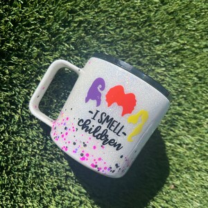 May include: White glitter mug with a black rim and handle. The mug has a purple, orange, and yellow silhouette of three witches with the text "I smell children" printed on the front.