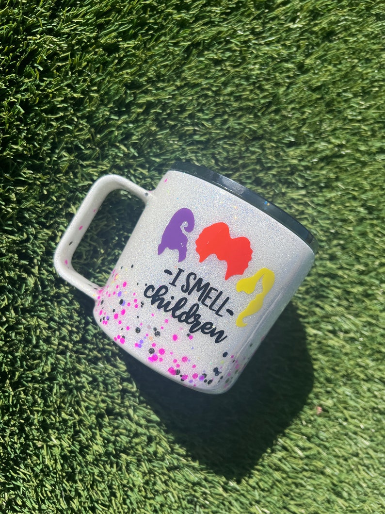 May include: White glitter mug with a black rim and handle. The mug has a purple, orange, and yellow silhouette of three witches with the text "I smell children" printed on the front.
