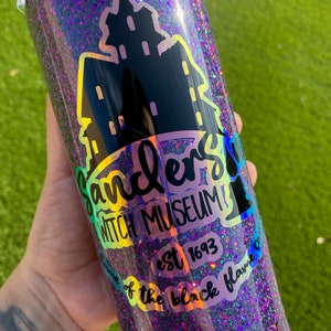 Puede incluir: Un vaso de glitter negro y morado con una silueta negra y blanca de una casa y el texto "Sanders Witch Museum est 1693 Home of the black flame c".