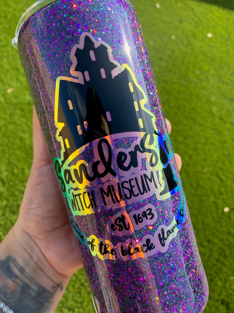 Puede incluir: Un vaso de glitter negro y morado con una silueta negra y blanca de una casa y el texto "Sanders Witch Museum est 1693 Home of the black flame c".