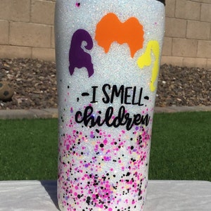 May include: White glitter tumbler with a black lid and a purple, orange, and yellow silhouette of the Sanderson sisters from Hocus Pocus. The text "I smell children" is printed on the tumbler.