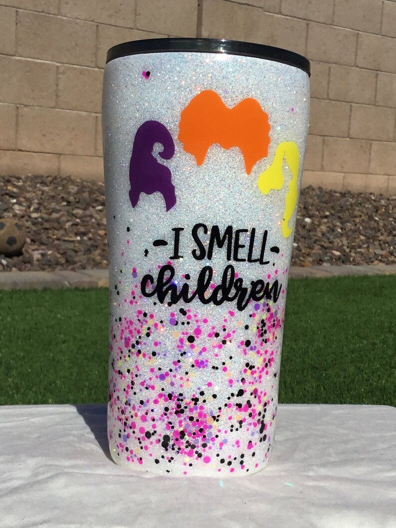 May include: White glitter tumbler with a black lid and a purple, orange, and yellow silhouette of the Sanderson sisters from Hocus Pocus. The text "I smell children" is printed on the tumbler.