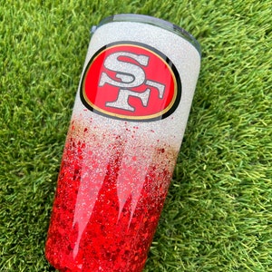 Może przedstawiać: Biały i czerwony kubek z brokatem z czerwono-złotym logo San Francisco 49ers.