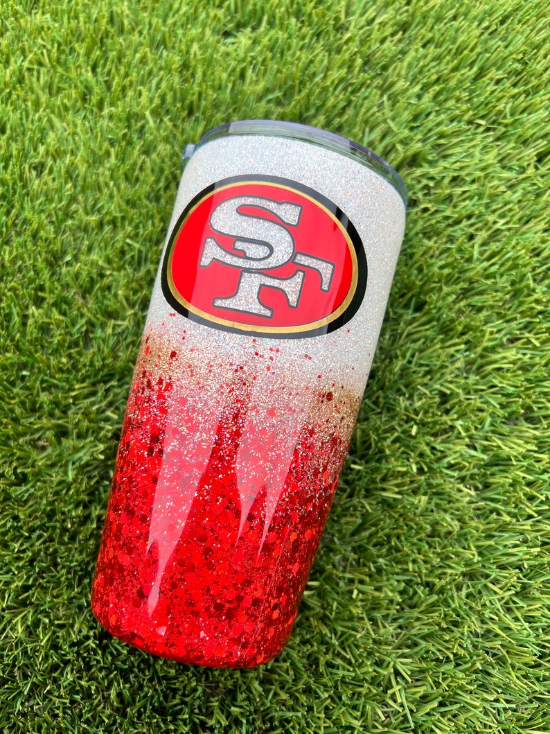 Może przedstawiać: Biały i czerwony kubek z brokatem z czerwono-złotym logo San Francisco 49ers.