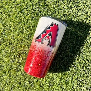 May include: A red and white glitter tumbler with a red and black Arizona Diamondbacks logo. The tumbler is on a green grassy surface.