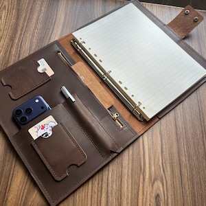 May include: A dark brown leather portfolio organizer featuring a six-ring metal binder with lined paper, a zippered pocket, a pen loop, and multiple slots for cards and a smartphone. The interior includes a snap-button closure strap.