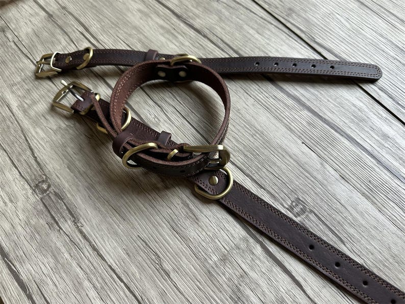 May include: Two brown leather dog collars with brass hardware. The collars are on a wooden surface.