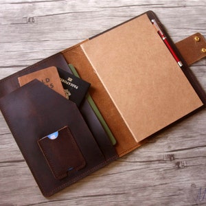 May include: A brown leather notebook cover with a pen holder, a pocket for a passport, and a smaller pocket for cards. The cover is open to reveal a blank notebook.