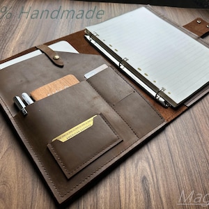 Può includere: Una cartella portadocumenti in pelle marrone con un raccoglitore ad anelli in metallo con carta a righe, una custodia per tablet con chiusura a scatto, diversi scomparti per carte e passanti per penne. Include una carta di credito dorata e il testo 100% Handmade e Magoda.