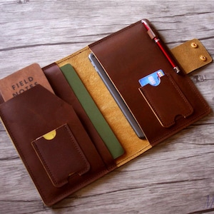 May include: A brown leather travel wallet with multiple pockets and slots for carrying cards, cash, and a pen. The wallet has a flap with two brass rivets and a pocket for a phone or tablet.