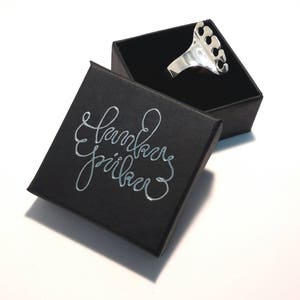 Puede incluir: Un anillo de plata con un diseño único que presenta cinco puntas. El anillo se muestra en una caja negra con las palabras "Joyería hecha a mano" impresas en letras plateadas.
