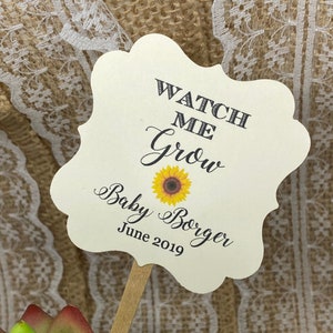 May include: A cream-colored paper sign on a wooden stick, with the text "WATCH ME Grow" and "Baby Borger June 2019" in black script. A yellow sunflower graphic is in the center. The sign is placed in front of a green succulent plant.