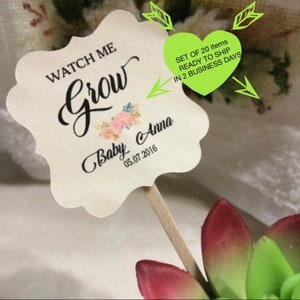 May include: A cream-colored, scalloped-edge tag on a wooden stick with the words "WATCH ME Grow" in black script. Below, it reads "Baby Anna 05.07.2016" with floral accents. A green heart-shaped graphic is also present.