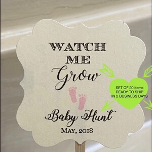 May include: A cream-colored, scalloped-edge sign on a wooden stick. The sign reads "WATCH ME Grow Baby Hunt MAY, 2018" in black script and block letters, with pink baby footprints. The sign is for a baby shower.