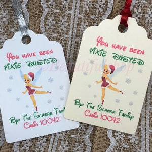 May include: Two white and cream-colored gift tags with a cartoon image of Tinkerbell in a red dress and a Santa hat. The text on the tags reads "You have been pixie dusted" and "By: The Schorr Family Cabin 10042".