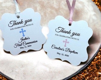 May include: Two white thank you tags with a cross design. The tag on the left has a blue cross and says "Thank you for celebrating with us Justin's First Communion May 11, 2024". The tag on the right has a pink cross and says "Thank you for celebrating with us Caroline's Baptism May 15, 2020".