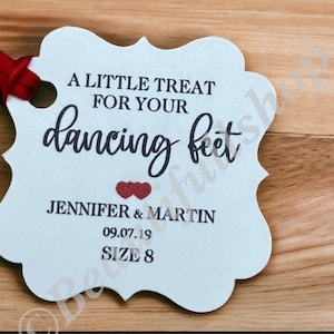 May include: A light blue, scalloped-edge tag with red ribbon. Text reads "A LITTLE TREAT FOR YOUR dancing feet" with names Jennifer & Martin, date 09.07.19, and size 8. The tag is on a wooden surface.