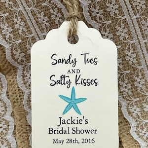 May include: A cream-colored tag with a scalloped edge, tied with twine. The tag features the text "Sandy Toes and Salty Kisses" with a turquoise starfish graphic. Below, it reads "Jackie's Bridal Shower, May 28th, 2016."