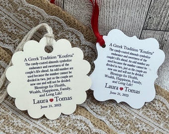 May include: Two white favor tags with a scalloped edge and a red heart. The tags feature the text "A Greek Tradition "Koufeta"" and explain the tradition of giving candy-coated almonds at weddings. The tags are personalized with the names "Laura" and "Tomas" and the date "June 21, 2023".