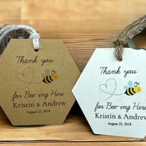 May include: Two hexagonal thank you tags with a bee and heart design. The text on the tags reads "Thank you for Bee-ing Here Kristin & Andrew August 25, 2018".
