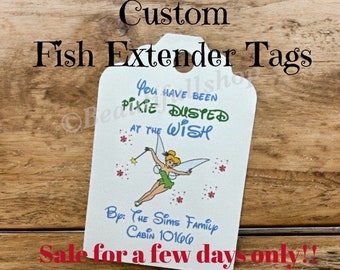 May include: A white tag with a green fairy illustration and the text "You have been pixie dusted at the wish" and "By: The Sims Family Cabin 10166". The tag has a hole at the top for hanging. The tag is on a wooden surface.
