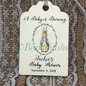 May include: Cream-colored gift tag with the text "A Baby is Brewing" and "Jackie's Baby Shower, September 9, 2018." Features a watercolor illustration of a rabbit in a blue jacket within a wreath. A gray string is attached.