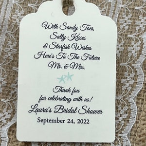 May include: A white tag with a scalloped edge and a brown twine bow. The tag has black text that reads "With Sandy Toes, Salty Kisses & Starfish Wishes Here's To The Future Mr. & Mrs. Thank you for celebrating with us! Laura's Bridal Shower September 24, 2022".