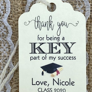May include: A white thank you tag with black text that reads "thank you for being a KEY part of my success."  The tag also features a black graduation cap and diploma, and the text "Love, Nicole CLASS 2020".