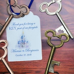 May include: Four antique-style key bottle openers with Mickey Mouse ears. The tag reads "Thank you for being a KEY part of our fairytale" with a watercolor illustration of Cinderella's castle. The tag also includes the names "Marisa & Benjamin" and the date "October 27, 2018".
