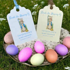 May include: Two light blue and cream colored baby shower favor tags with a Peter Rabbit illustration. The tags say "Thanks for Hopping by Jacqueline's Baby Shower September 9, 2016". The tags are sitting on a silver tray with pastel colored eggs.