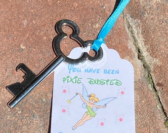 May include: A silver key with a Mickey Mouse-shaped bow is attached to a white tag with a blue ribbon. The tag features the text "You have been Pixie Dusted" and an illustration of Tinkerbell.